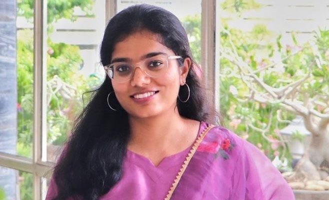 Dr Naazia Siddiqua standing indoors wearing a purple floral-patterned saree, with long dark hair and hoop earrings, positioned in front of large windows with greenery visible outside.
