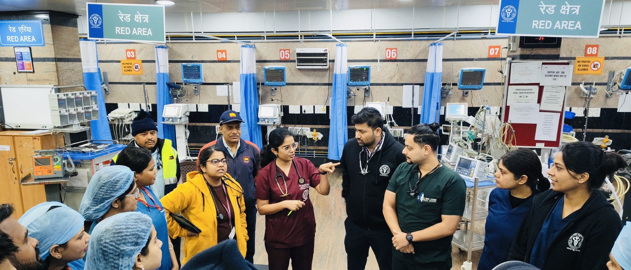 Emergency Medicine clinician leading a team briefing in the resuscitation area, with staff listening and equipment visible in the background.