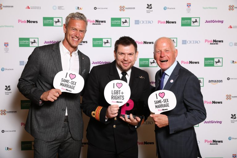 Three men in business suits, photographed at a Pink News event holding "I Love Pink News" signs.