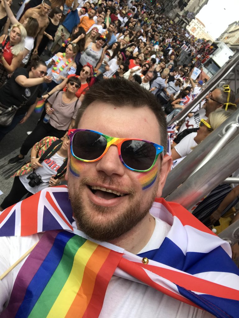 Mike Hill is leaning out of a bus with a crowd behind him. He is wearing a British and Pride flag around their chest.