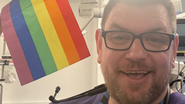 Mike Hill in a hospital cubicle holding a rainbow flag.